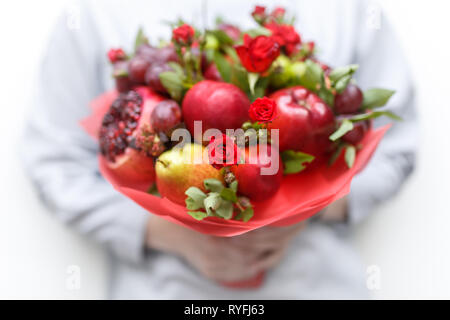 Genießbare Blumenstrauß aus Granatapfel, Äpfel, Pflaumen und roten Rosen in die Hände der Frau auf weißem Hintergrund Stockfoto