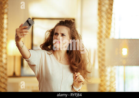 Junge Hausfrau mit langen brünetten Haar in der modernen Wohnzimmer Fang ein schwaches WLAN-Signal auf dem Smartphone.