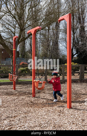 Zwei Jahre alte Junge spielt mit Schaukeln im Park trägt einen Bobble hat und Red Coat Stockfoto