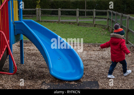 Zwei Jahre alten Jungen spielen auf dem Kunststoff Folie im Park trägt einen roten Mantel und Bobble hat Stockfoto