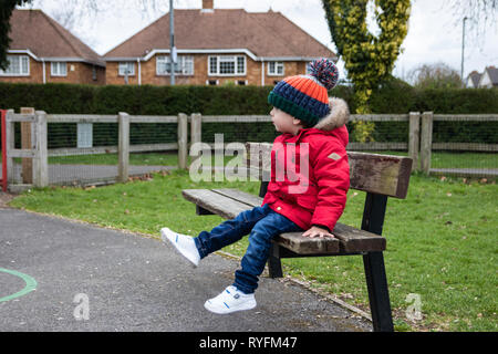 Zwei Jahre altes Kleinkind saß auf einer Parkbank seine Beine schwingen, trägt einen roten Mantel und Bobble hat Stockfoto