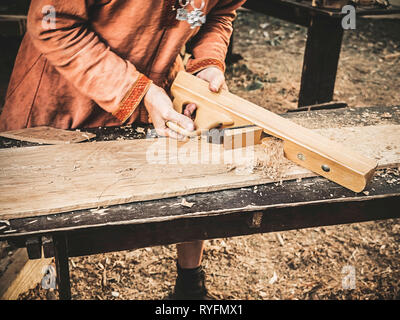 Tischler in mittelalterlicher Kleidung aus Baumwolle, mit einem Holz mit dem Flugzeug. Man Manuell gießt Sägemehl aus der Planer. Bau eines hölzernen Barke Stockfoto