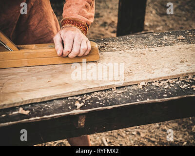 Tischler in mittelalterlicher Kleidung aus Baumwolle, mit einem Holz mit dem Flugzeug. Man manuell Hews ein Holzbrett mit einem Flugzeug. Bau eines hölzernen Barke Stockfoto
