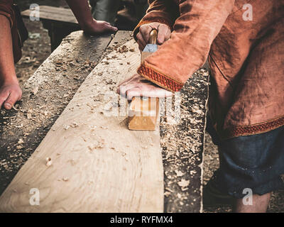 Tischler in mittelalterlicher Kleidung aus Baumwolle, mit einem Holz mit dem Flugzeug. Man manuell Hews ein Holzbrett mit einem Flugzeug. Bau eines hölzernen Barke Stockfoto