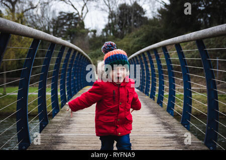 Kleiner Junge auf die Kamera läuft auf Holzbrücke schreien und Spaß haben, trägt einen roten Mantel und Bobble hat Stockfoto