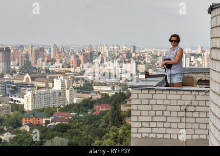 Weiß im mittleren Alter gegerbt Frau steht auf dem Dach eines hohen Gebäudes mit Antenne Stadtbild in Kiew, Ukraine. Stockfoto