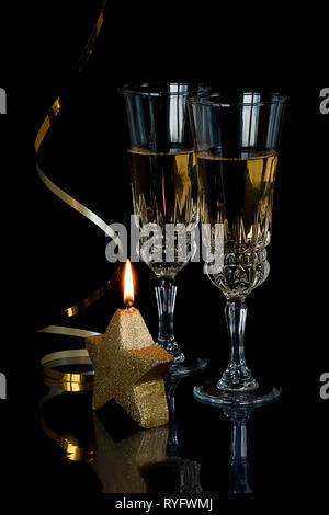 Festliche Zusammensetzung: Zwei Kristallgläser mit Champagner, golden Serpentine und eine brennende Kerze in der Form eines Sterns sind auf schwarzem Hintergrund Stockfoto