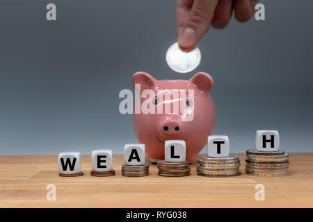 Symbol für zunehmenden Wohlstand. Würfel auf Stapel von Münzen Form das Wort "Reichtum". Im Hintergrund eine Münze in ein Sparschwein. Stockfoto