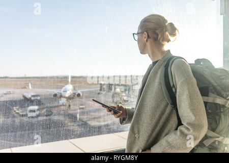 Leger gekleidete weibliche Reisende am Flughafen, Smart Phone, schauen durch den Flughafen Tor Fenster Ebenen auf Landebahn Stockfoto