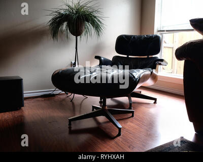 Charles und Ray Eames Designer schwarz Lounge Sessel mit Ottomane in einem Wohnzimmer Einstellung durch ein Fenster auf einen Holzboden, gedämpften Tönen und dunkel". Stockfoto