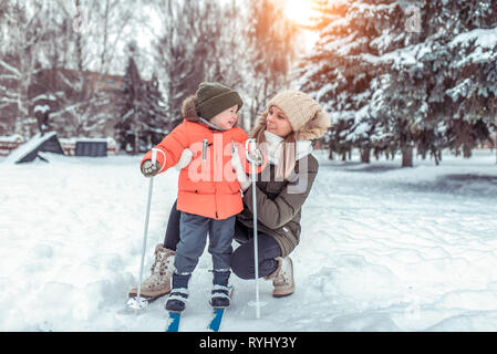 Eine junge Frau, die Mutter unterstützt mit Sorgfalt, kleinen jungen Sohn 3-6 Jahre alt, Skifahren im Winter Forest Park. Der erste Sport Unterricht auf der Straße. Glücklich Stockfoto