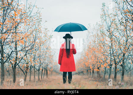 Frau im roten Mantel und mit Regenschirm zwischen Bäumen in Apple Garten im Herbst. Minimalismus, Reisen, Natur Konzept. Stockfoto