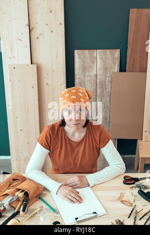 Portrait von selbständigen weiblichen Tischler tragen bandanna am Schreibtisch in Ihrem Holzarbeiten Workshop durch verschiedene Tools des Handels umgeben Sitzen Stockfoto