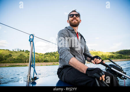 Ein Mann mit Sonnenbrille und legere Kleidung, als er ein kleines Beiboot rund um einen See oder Fluss treibt. Stockfoto