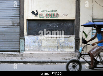Eine Fahrradrikscha (Fahrrad Taxi) Fahrten entlang einer Straße in der Altstadt von Havanna, Kuba, vorbei an einem geschlossenen Einzelhandel Fenster mit einer spanischen Schild El Gallo (der Hahn). Stockfoto