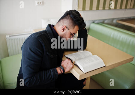 Arabischer mann Verschleiß auf schwarze Jeans Jacke und Brille im Cafe sitzen, Buch lesen. Elegantes und Modisches arabischen Modell Kerl. Stockfoto