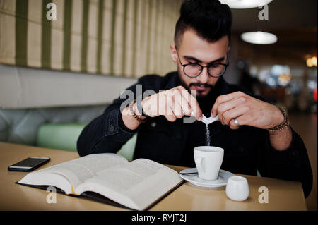 Arabischer mann Verschleiß auf schwarze Jeans Jacke und Brille im Cafe sitzen und Kaffee trinken, Buch lesen. Elegantes und Modisches arabischen Modell Kerl. Gießen Sie Zucker