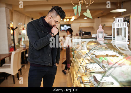 Arabischer mann Verschleiß auf schwarze Jeans Jacke und Brillen in Cafe wählen Sie Kuchen. Elegantes und Modisches arabischen Modell Kerl. Stockfoto