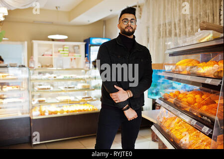 Arabischer mann Verschleiß auf schwarze Jeans Jacke und Brillen in Cafe wählen Bäckerei. Elegantes und Modisches arabischen Modell Kerl. Stockfoto