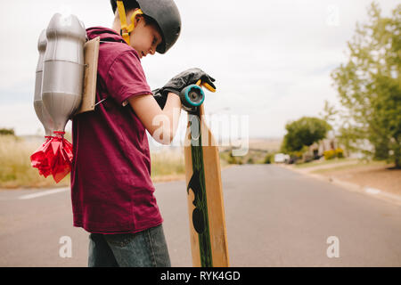 Junge mit Helm mit einem Spielzeug Jetpack auf dem Rücken stehen im Freien mit seinem Skateboard. Kind mit Jetpack und Skateboard. Stockfoto