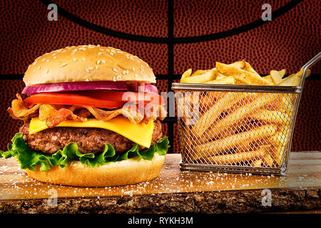 Cheeseburger mit Speck und einen Korb mit Pommes frites. Basketball Ball Bild im Hintergrund. Stockfoto