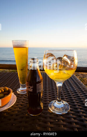 Glas Bier und Whiskey neben einem Coca Cola auf der Terrasse mit Mittelmeer im Hintergrund. Spanien. Stockfoto