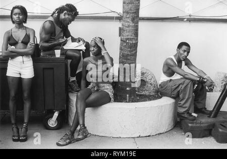 South Beach, Miami, Florida, USA, 1990er Jahre Eine Gruppe junger afroamerikanischer Männer und Frauen, die in Shorts und T-Shirts gekleidet sind, sitzen und warten, ganz in ihren eigenen Gedanken versunken. Ein Mann macht Bucharbeit, während eine andere Frau eine Weinflasche in einem Papierbeutel zwischen ihren Beinen hält. 1999 USA HOMER SYKES Stockfoto