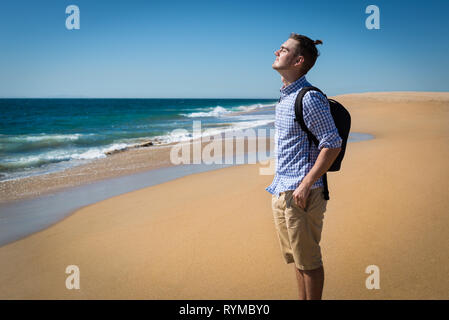 Glückliche junge Mann im blauen Hemd mit Rucksack mit sonnigen Sommertag am Strand in Kalifornien, USA. Hübscher Kerl alleine stehen mit Blick auf den Ozean Stockfoto