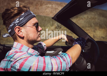 Stilvolle Mann in Kopfbügel und bunten T-Shirt fahren Cabrio Auto, Seitenansicht. Attraktiver Kerl mit Road Trip im Sommer sonnigen Tag in Kalifornien, USA. Stockfoto