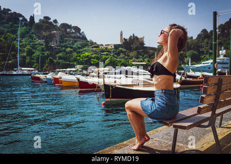 Jungen barfuß Frau sitzt auf der Bank in der Marina in Portofino, Italien. Weibliche Touristen genießen sonnigen Tag auf dem Hintergrund der kleinen Dorf, Meer und Boote. Stockfoto