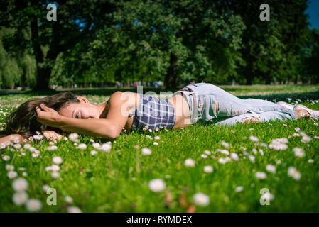 Junge fröhliche Frau liegt auf grünem Gras. Happy girl ist entspannend im Kamille Feld in warmen Sommertag auf dem Hintergrund der großen Bäume im Park. Stockfoto