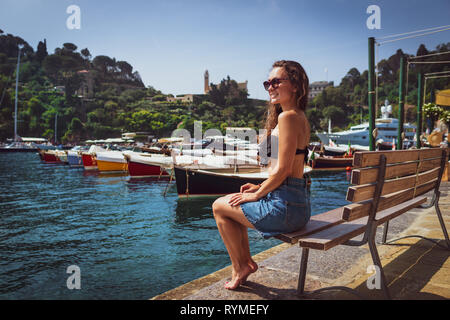 Jungen barfuß Frau auf der Bank in der Marina in Portofino, Italien sitzen. Weibliche Touristen genießen sonnigen Tag auf dem Hintergrund der kleinen Dorf, Meer und Boote. Stockfoto