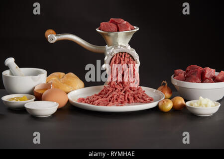 Fleischwolf mit Schweinefleisch oder Rindfleisch auf Küche mit Zutaten zum Kochen Schalen in Weiß auf schwarzem Hintergrund Stockfoto
