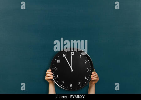 Mann hält runde Büro Uhr zeigt 5 Minuten zu Mittag am Schwarzen Brett Hintergrund Stockfoto