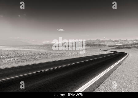 Gepflasterte Straße durch die karge Wüste in Richtung Berge in der Ferne geschwungen. Schwarz und Weiß. Stockfoto