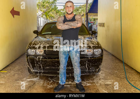 Eine stolze junge Mann steht vor seinem Seifigen Auto. Einen männlichen Mann mit Tattoos dauert einen Moment vor seinem Seifigen Auto innen die Waschhalle zu stellen.