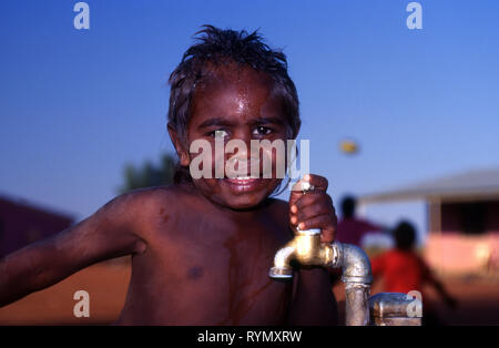 JUNGER LÄCHELNDER ABORIGINE-JUNGE, DER SICH AM ZAPFHAHN HÄLT, YUELAMU ABORIGINE COMMUNITY (MOUNT ALLAN SCHOOL) NORTHERN TERRITORY, AUSTRALIEN. Stockfoto