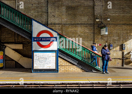 Eine Frau schaut auf ihr Mobiltelefon, während sie auf einen U-Bahn-Zug der Wimbledon-Linie an der U-Bahn-Station West Brompton, London, Großbritannien, wartet Stockfoto