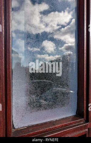 Single verglaste Tür fenster mit Kondenswasserbildung auf der Innenseite der Tür aus Glas mit Cloud Reflexionen in der frostigen Glas Stockfoto
