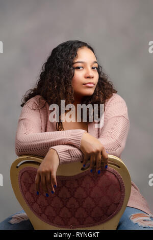 Man jugendlich. Schön, Biracial an der High School mit dem lockigen Haar in den Stuhl nach hinten mit den Armen nachdenklich überquert an der Kamera schaut. Stockfoto