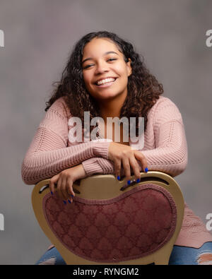 Eine attraktive weibliche biracial jugendlich nach hinten sitzen im Stuhl lächelnd mit perfekte Zähne für ältere Portraits posiert. Stockfoto