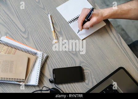 Der Mann hält einen Stift und schreibt in einem Notebook. In der Nähe befinden sich das Smartphone, Kopfhörer, Computer. Stockfoto