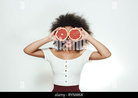 Gesunde Lebensweise. Verspielte junge afro-amerikanische Frau, die zwei Teile der Grapefruit auf ihre Augen und lächelte, während gegen grauen Hintergrund stehen. Früchte. Gesunde Ernährung Konzept Stockfoto
