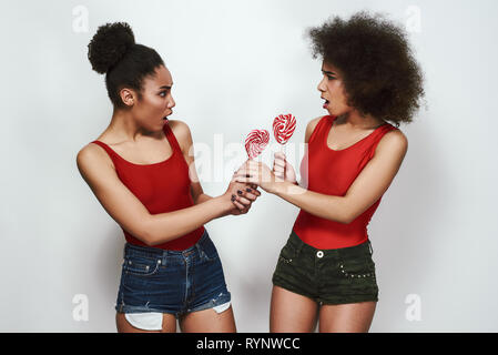 Ich will mehr! Zwei verärgerte afro-amerikanische Frau, die in Jeans Shorts sind sich einander mit geöffnetem Mund und Holding Lutscher geformte Herz, während gegen grauen Hintergrund stehend Stockfoto