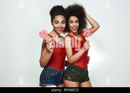 Süsse Schönheit. Zwei schöne junge afro-amerikanische Frauen im Sommer Kleidung holding Lutscher geformte Herzen und lächelnd, während gegen grauen Hintergrund stehend Stockfoto