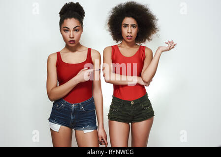 Es ist Ihr Fehler! Studio shot von enttäuscht afro-amerikanische Frau, die in Jeans Shorts zeigen Zeigefinger zu ihrer Freundin, während gegen grauen Hintergrund stehen. Wird verrückt. Negative Emotionen Stockfoto