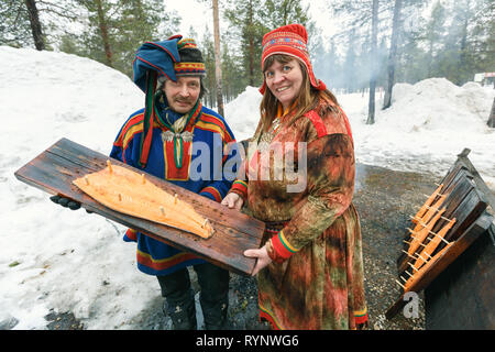 Saariselka, Finnland - 24 April 2013: Lokale samische Paar hält Holzbrett mit geräuchertem Lachs filet auf Open air Feuer in Saariselka, Fin vorbereitet Stockfoto