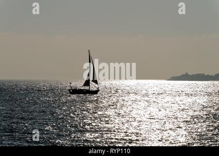 Silhouette einer Yacht segeln auf den Solent in Portsmouth Hampshire, gegen die Reflexion von Sonnenlicht auf dem Meer, England Großbritannien Stockfoto