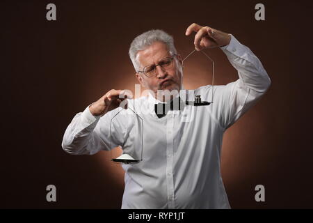 Ältere grauhaarige Mann 50s, in weißem Hemd, Gläser und Bow Tie etwas Wiegen auf Skalen, die mit kleinen Kettlebells Stockfoto