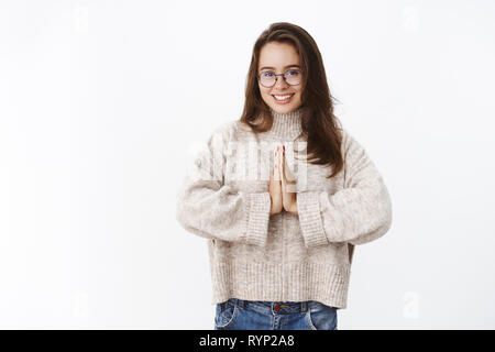 Studio geschossen von süß und weibliche Junge 20 s Frau in Gläsern und Pullover holding Handflächen zusammen, die sich in der Nähe des Körpers in namaste Geste breit grinsend Gruß Stockfoto